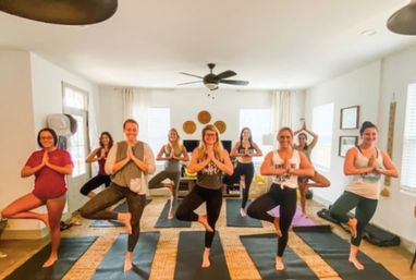Bright indoor yoga studio: a group of women in a community class holding tree pose on yoga mats under a ceiling fan and woven wall decor.