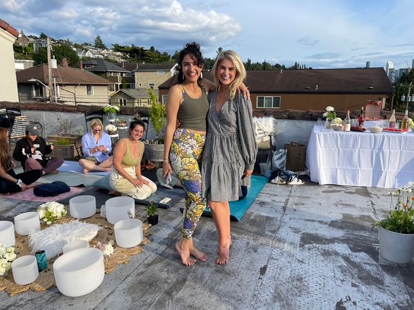 Two smiling women barefoot pose on an urban rooftop during a daytime wellness gathering, surrounded by yoga mats, white singing bowls, cushions, flowers and a drinks table, with houses and a city skyline in the background.