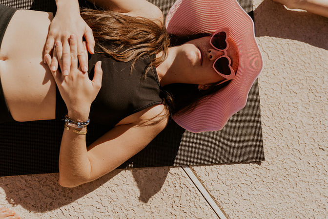 Woman sunbathing on a black mat on a sunny patio, wearing a pink floppy hat and heart-shaped sunglasses with hands folded on her stomach.
