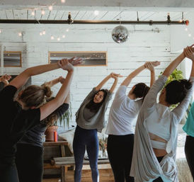 Group yoga/stretching class in a bright loft-style wellness studio, participants in activewear reaching arms overhead beneath string lights and a disco ball, with potted plants and wooden benches.