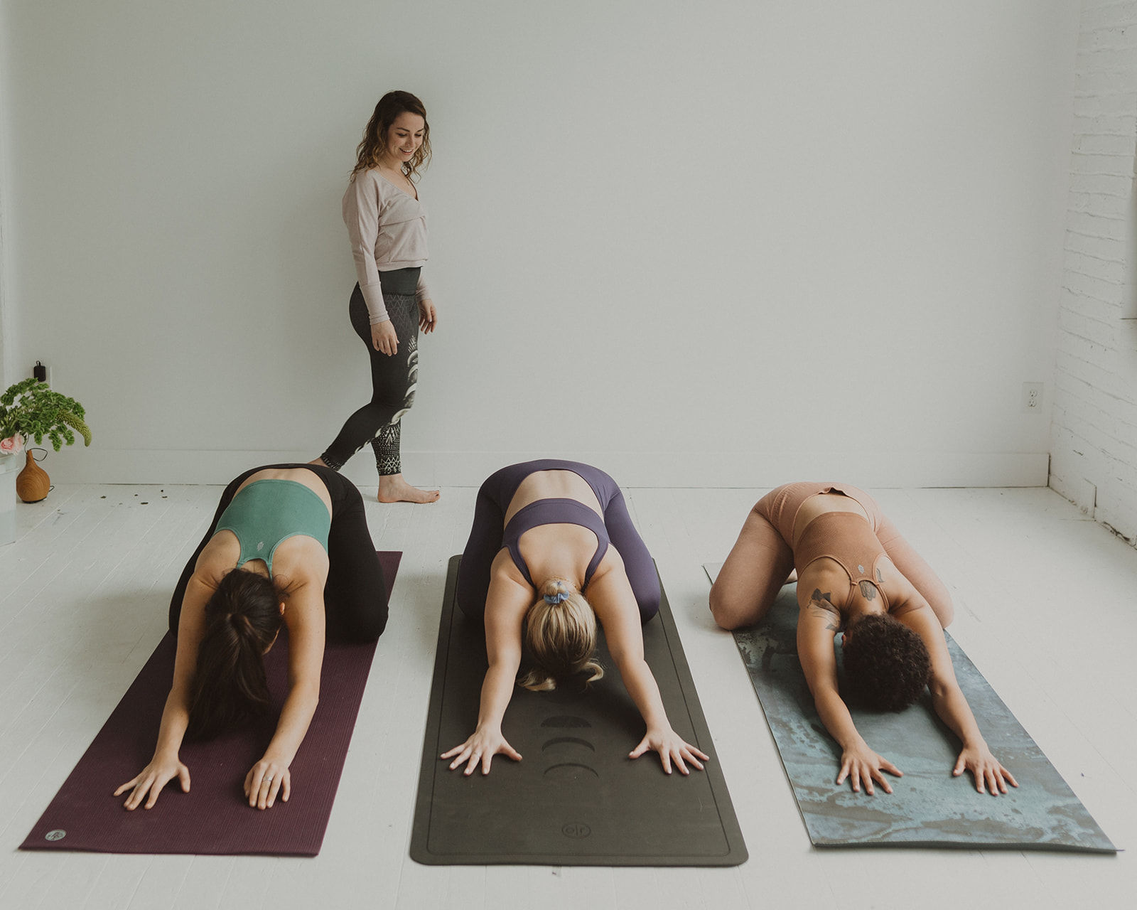 Calm group yoga class in a bright minimalist studio — instructor watching three students on mats practicing child's pose.