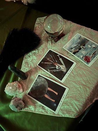 Moody divination scene on shimmery satin: crystal ball on a wooden stand, three tarot cards (one showing swords imagery), cluster crystals and a black feather fan bathed in green light.