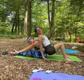 Partner yoga in a leafy forest: two friends on green and purple mats doing a seated back-to-back stretch on wood-chip ground, smiling beneath tall trees.