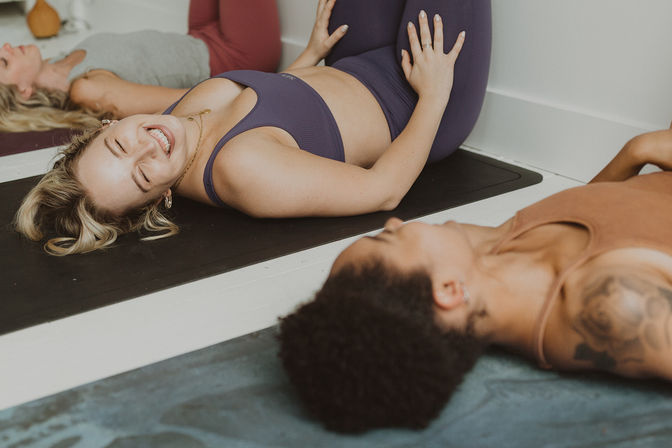 Smiling woman in purple activewear doing a knee-to-chest stretch on a yoga mat during a relaxed indoor group yoga class with others in a bright fitness studio