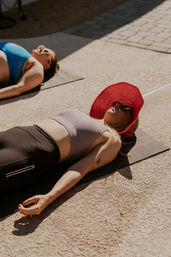 Two women sunbathing on yoga mats on a sunny poolside patio — foreground woman wearing a red wide-brim hat and sunglasses, gray crop top and black leggings, relaxed with painted nails.