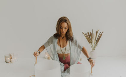 Person seated cross-legged in a bright wellness studio playing two large crystal singing bowls with mallets, surrounded by lit candles and dried pampas grass — sound healing meditation session
