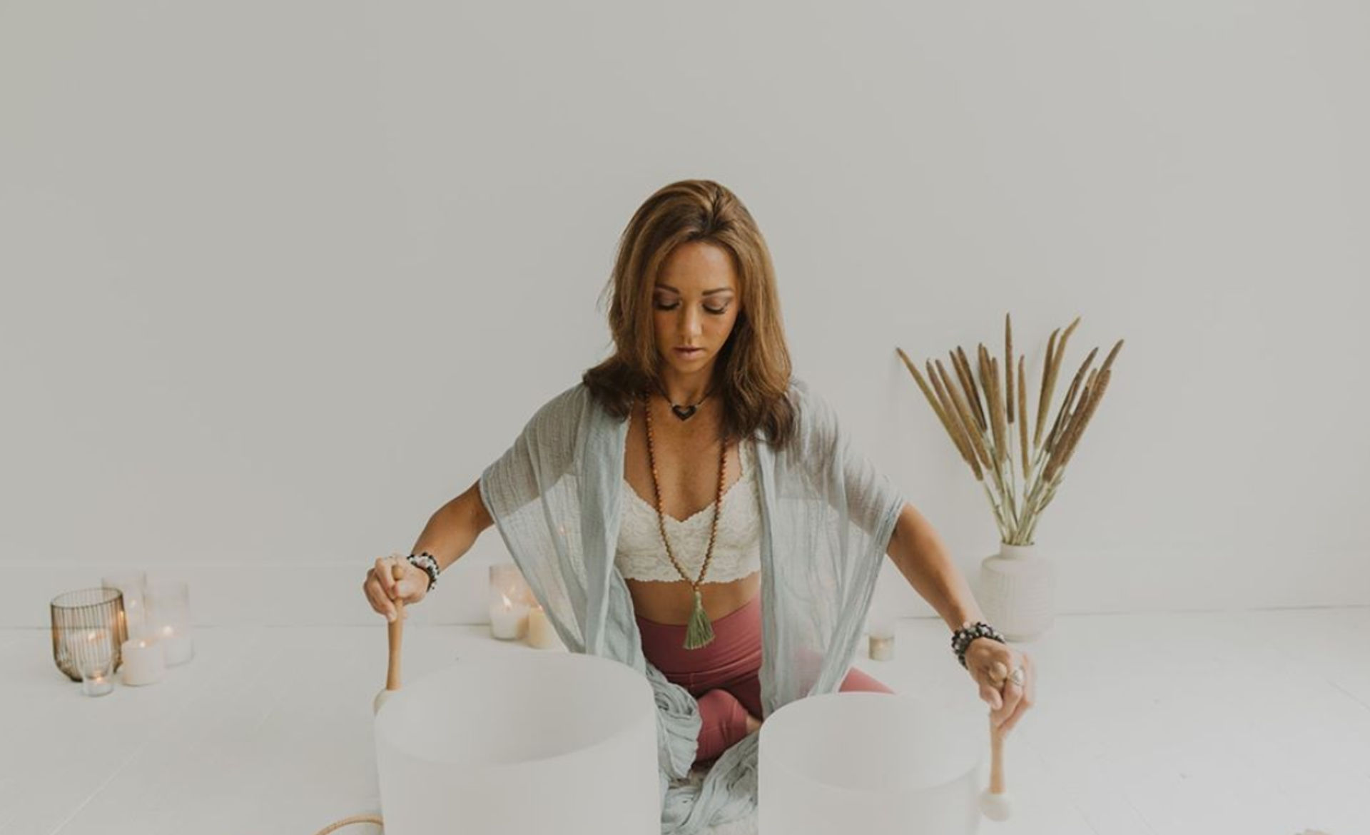 Person seated cross-legged in a bright wellness studio playing two large crystal singing bowls with mallets, surrounded by lit candles and dried pampas grass — sound healing meditation session