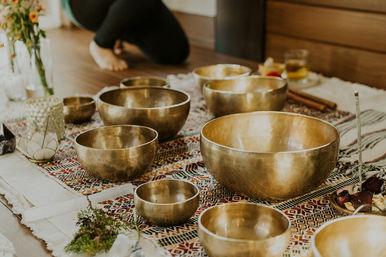Brass singing bowls arranged on a colorful woven mat for a sound bath in a cozy meditation space, with incense, candles, crystals and a kneeling participant in the background.
