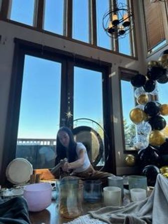Sound-bath facilitator seated by a gong and crystal singing bowls in a sunlit wellness studio with tall windows, black-and-gold balloons, and a bright city view beyond.
