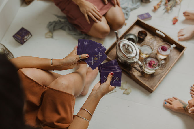 Hands holding purple tarot cards in a cozy indoor tarot reading beside a wooden tray with a glass teapot and floral tea cups on a white floor, people seated cross‑legged.