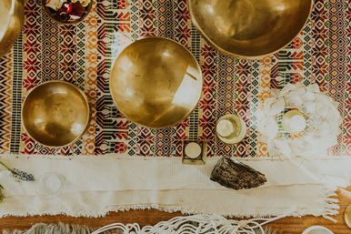 Top-down view of a cozy boho meditation altar: brass singing bowls, candles, a smudge stick and colorful woven rug.