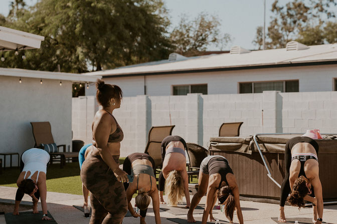 Outdoor group yoga class on a sunny residential patio, instructor guiding participants in forward-fold poses on mats near patio furniture and a hot tub
