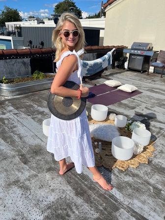 Smiling barefoot woman in a white sundress and sunglasses on an urban rooftop holding a gong beside crystal singing bowls arranged on a woven mat for a rooftop sound-bath/yoga session, with a yoga mat, pillows, grill and low-rise buildings under a sunny blue sky.