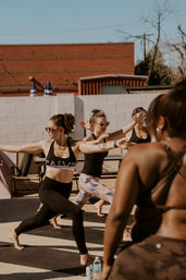 Sunlit outdoor urban yoga class with women on mats doing warrior pose in a brick courtyard, some wearing playful heart‑shaped sunglasses and water bottles nearby.