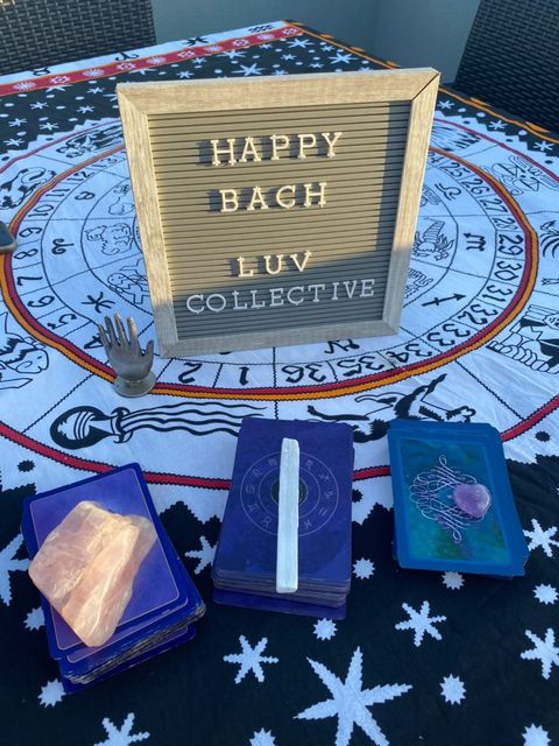 Playful outdoor tarot setup on a zodiac tapestry: wooden letterboard reading 'HAPPY BACH LUV COLLECTIVE', three card decks, selenite wand, rose quartz crystal and a small decorative hand on a patio table.