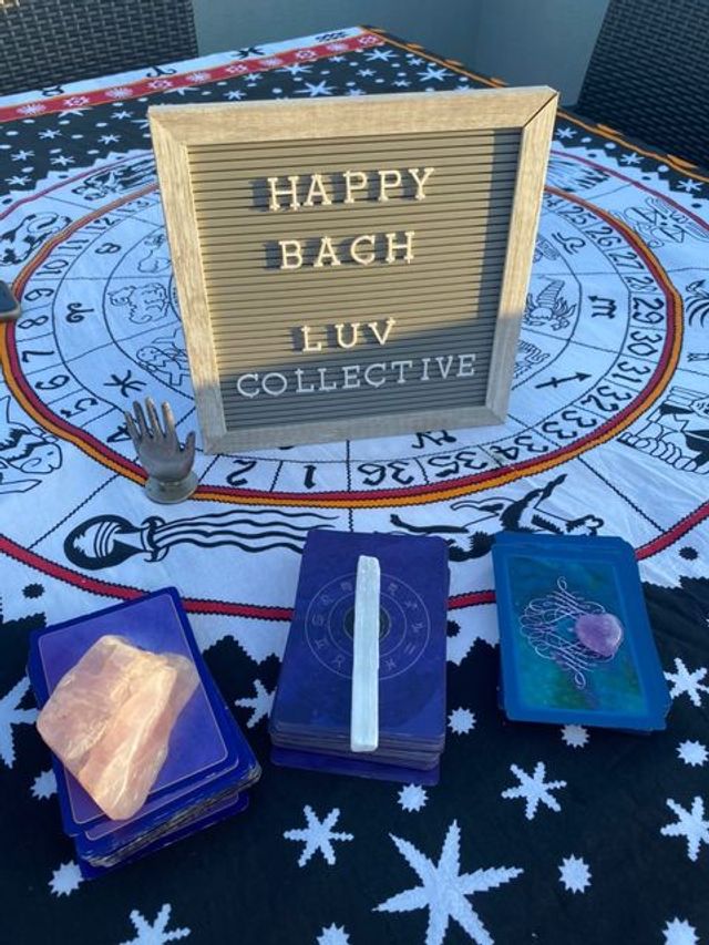 Playful outdoor tarot setup on a zodiac tapestry: wooden letterboard reading 'HAPPY BACH LUV COLLECTIVE', three card decks, selenite wand, rose quartz crystal and a small decorative hand on a patio table.