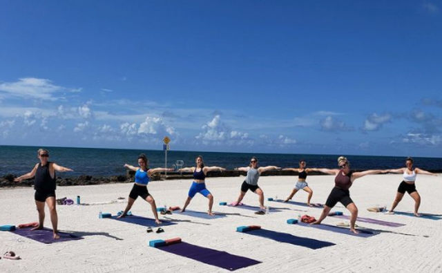 Seven people practicing beach yoga on colorful mats and blocks, striking Warrior II poses on a sunny sandy shore with calm blue ocean and sky