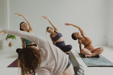 Three women in a bright yoga studio smiling and doing seated side stretches on mats during a guided group yoga class.