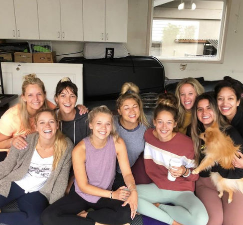 Smiling group of friends in casual activewear gathered in a bright home garage lounge, sitting on mats with coffee and a small fluffy dog.