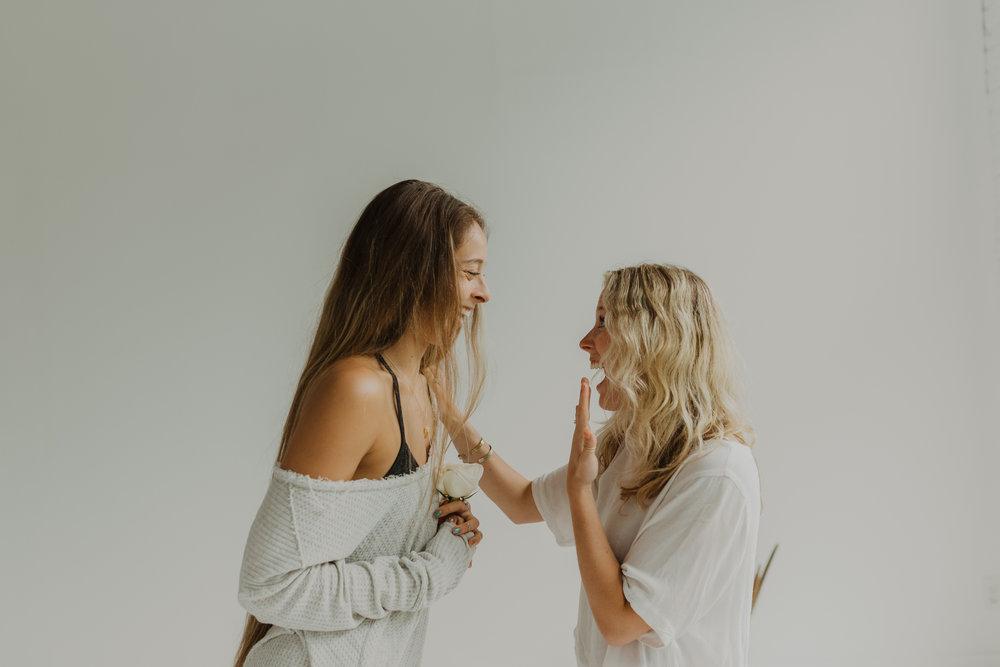 Two smiling women in a bright minimalist studio, one holding a white rose as they laugh and high-five