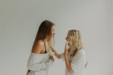Two smiling women in a bright minimalist studio, one holding a white rose as they laugh and high-five