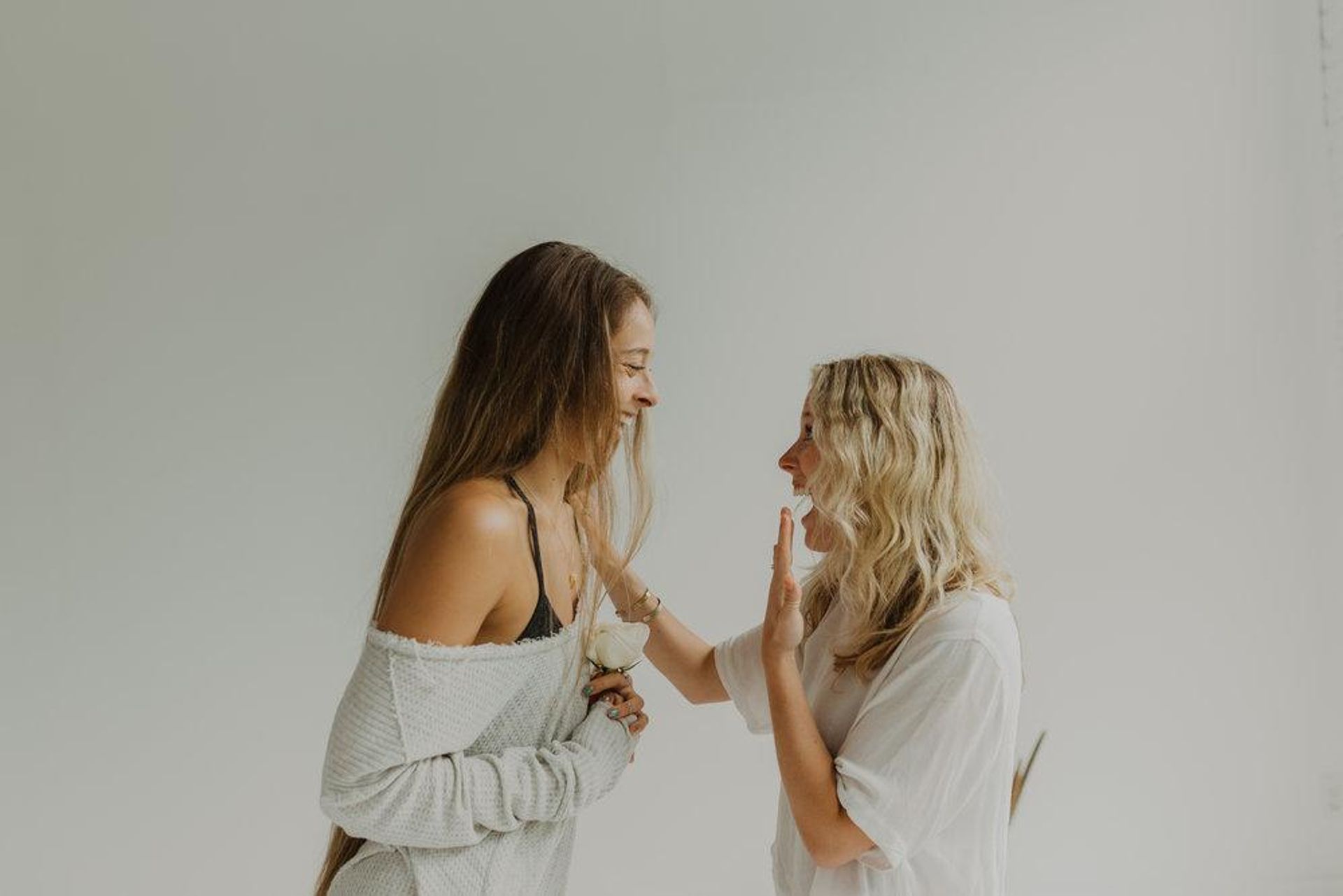 Bright minimalist studio portrait of two laughing women facing each other, one holding a white rose while the other raises a hand in playful surprise.