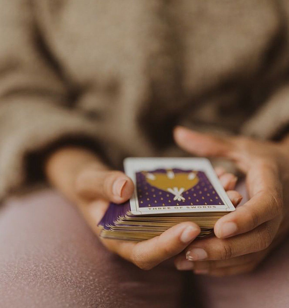 Close-up of hands holding a stacked tarot deck with the Three of Swords card face-up, cozy indoor tarot reading vibe.