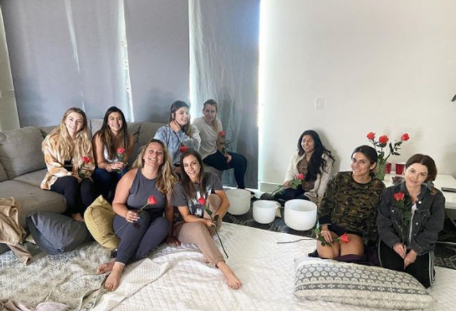 Group of nine women in a cozy living room circle, seated on a sofa and floor cushions holding red roses around white crystal singing bowls for a relaxed sound‑healing wellness gathering.