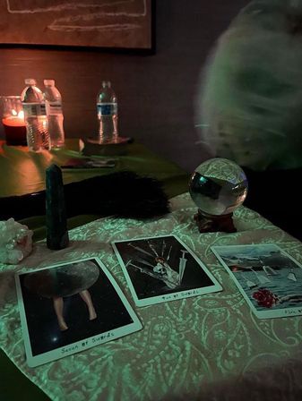 Dimly lit indoor tarot reading: three tarot cards spread on a patterned cloth, a crystal ball on a wooden stand, a green crystal obelisk and quartz cluster, a lit candle and water bottles in the background — moody, mystical vibe.