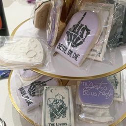 Tiered display of individually wrapped wedding-favor sugar cookies on a gold-edged stand: lavender “Bride or Die” tombstone with skeleton hand, white heart cookie with ring, “’Til Death Do Us Party” plaque and a tarot-style “The Lovers” skeleton couple.