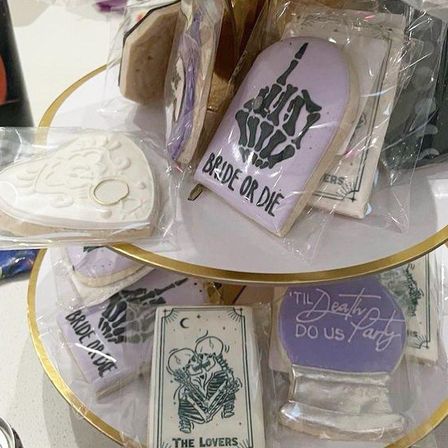 Tiered display of individually wrapped wedding-favor sugar cookies on a gold-edged stand: lavender “Bride or Die” tombstone with skeleton hand, white heart cookie with ring, “’Til Death Do Us Party” plaque and a tarot-style “The Lovers” skeleton couple.
