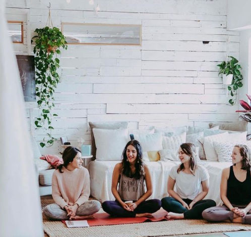Four women sitting cross-legged on yoga mats in a cozy, light-filled boho studio with white shiplap walls, hanging plants and cushions, smiling during a group yoga/meditation session