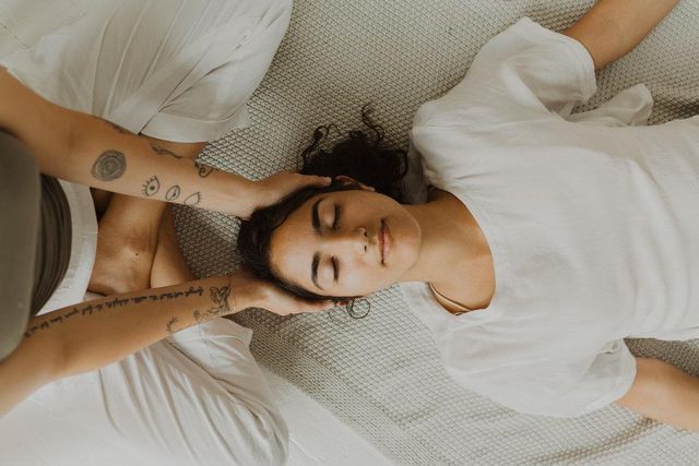Overhead shot of a woman lying on a textured mat with eyes closed as a tattooed practitioner gently cradles her head — calming indoor wellness/spa head massage scene in soft neutral tones.