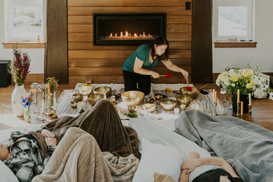 Woman leading an indoor sound bath with singing bowls in a cozy living room by a fireplace, participants lying under blankets amid flowers, candles and brass bowls.