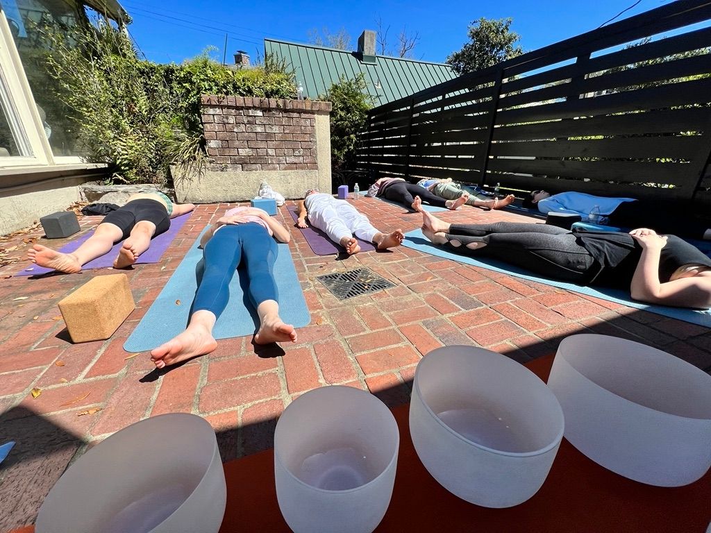 Outdoor yoga class on a sunny brick patio with students lying in savasana on colorful mats, crystal singing bowls in the foreground and a wooden fence and garden plants in the background.