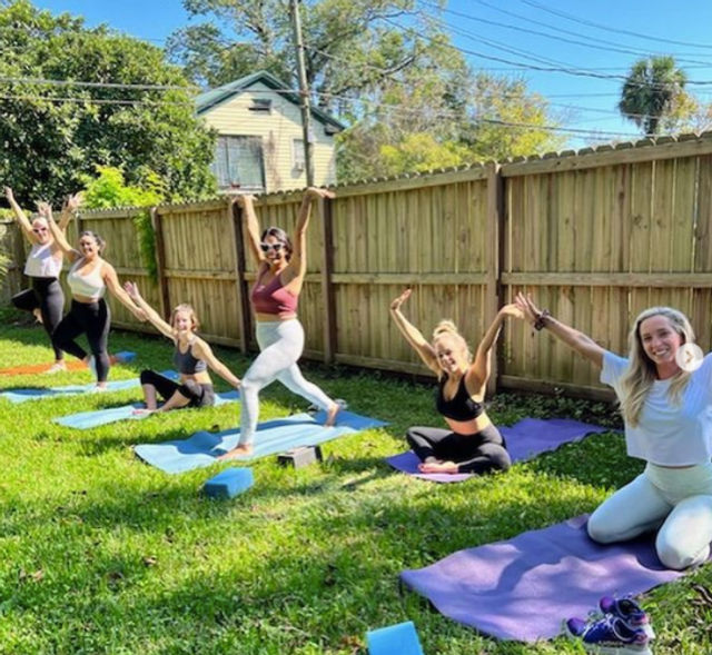 Group of six women practicing outdoor yoga on colorful mats in a sunny fenced backyard, smiling and striking energetic poses on a green lawn — outdoor group fitness and wellness scene.