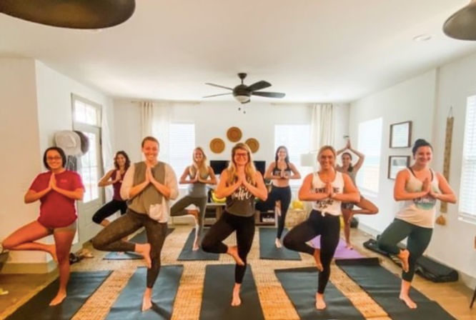 Bright home-studio yoga class, a group of participants practicing tree pose on mats in a living-room setting — casual activewear, barefoot, group fitness and wellness session.