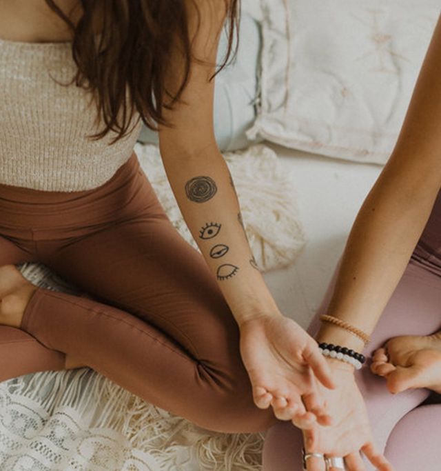 Two people seated cross-legged in a cozy boho space practicing yoga/meditation; close-up on a tattooed forearm with eye and swirl designs, beaded bracelets and earth-toned leggings.