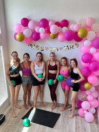 Six women in colorful athletic wear holding small exercise balls under a pink-and-gold balloon arch and “MISS TO MRS” banner in a bright indoor home workout studio with a yoga mat on a light wood floor