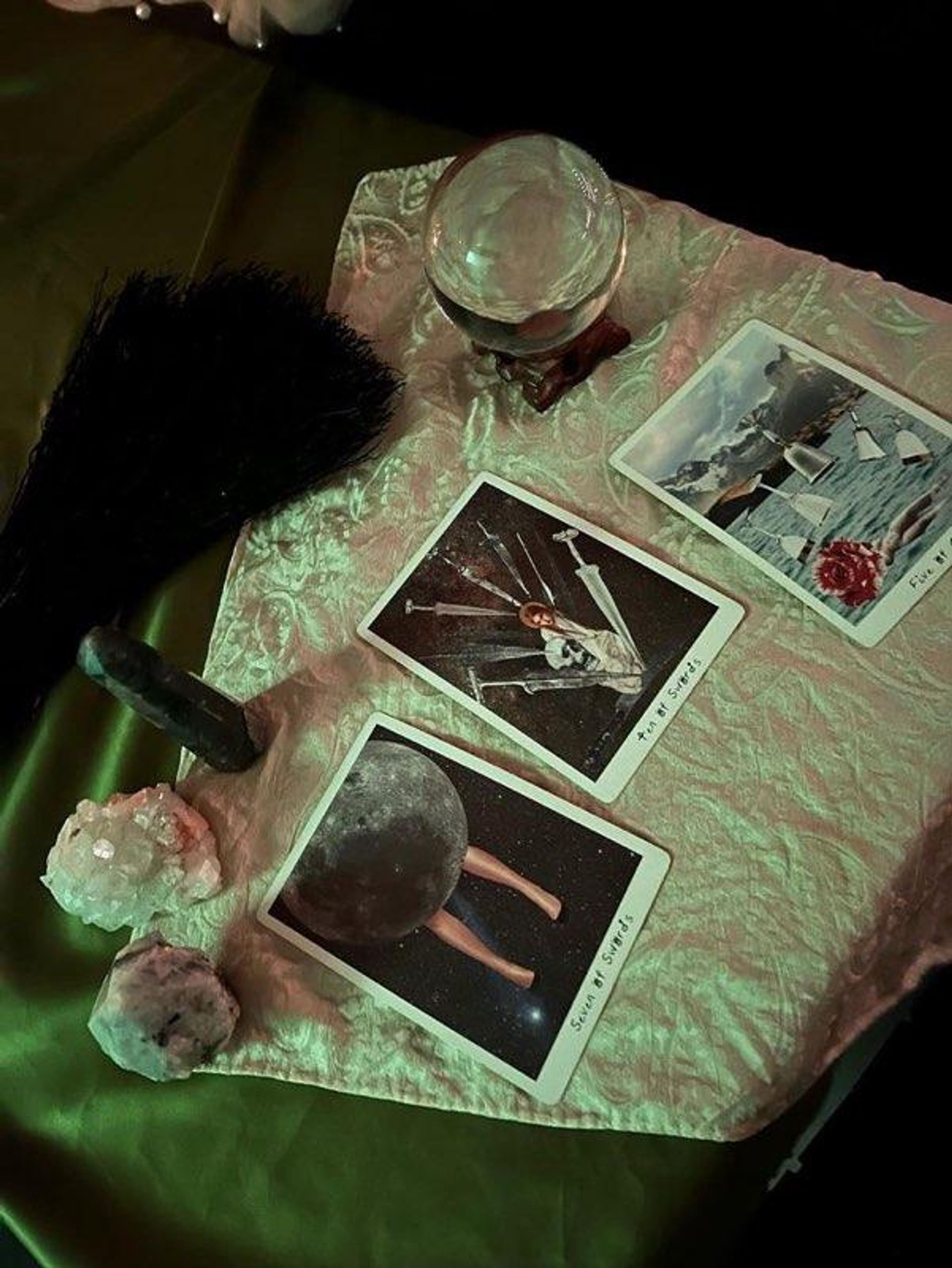 Mystic tarot reading table with three tarot cards on a satin cloth, a crystal ball, cluster crystals, a dark crystal point and a black feather fan under moody green lighting.