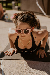 Person doing an outdoor yoga push-up on a black mat on a sunny patio, wearing pink heart-shaped sunglasses, a black sports top, gold chain, curly hair and a small bird tattoo on the shoulder.