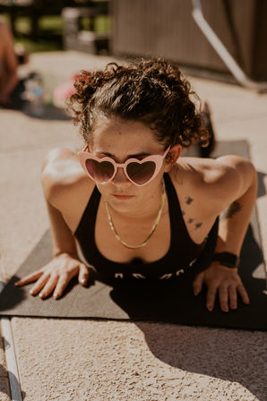Person doing an outdoor yoga push-up on a black mat on a sunny patio, wearing pink heart-shaped sunglasses, a black sports top, gold chain, curly hair and a small bird tattoo on the shoulder.