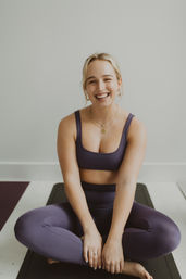 Smiling woman in purple activewear sitting cross‑legged on a black yoga mat in a bright indoor studio, wearing gold necklaces and hoop earrings
