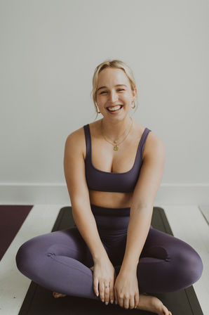 Smiling woman in purple activewear sitting cross‑legged on a black yoga mat in a bright indoor studio, wearing gold necklaces and hoop earrings