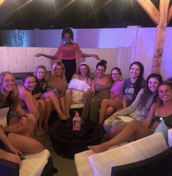 Dozen women smiling on a backyard patio lounge under string lights and a wooden pergola at night, seated around a round coffee table with a lantern and purple-hued party lighting — casual friends' night.