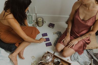 Two women on a white floor sharing a cozy indoor tarot reading with purple cards, candles, teapot, and boho cushions.