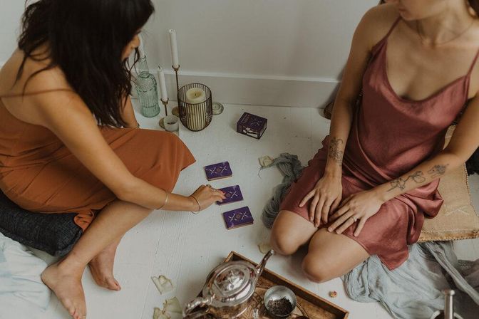 Two women on a white floor sharing a cozy indoor tarot reading with purple cards, candles, teapot, and boho cushions.