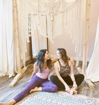 Two women in yoga clothes sitting on a patterned rug, smiling under a large boho macramé canopy in a cozy indoor yoga studio