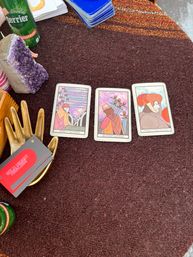 Three tarot cards (Nine of Swords, Seven of Swords, Page of Pentacles) laid out on a brown textured tablecloth with an amethyst crystal, gold hand card dish holding business cards, stacked decks and a canned sparkling drink — cozy outdoor tabletop tarot reading scene.