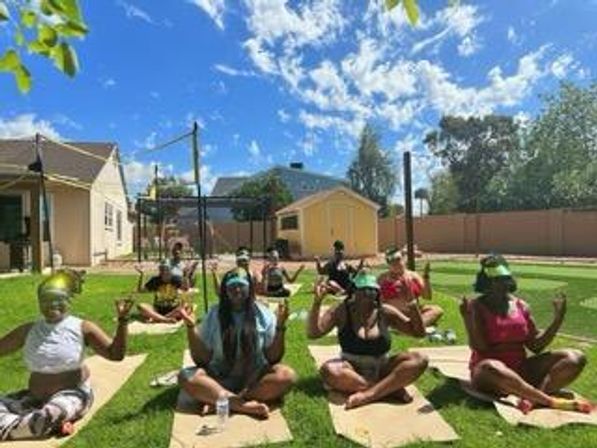 Sunny suburban backyard outdoor yoga class — several people seated cross-legged on mats in meditation pose, wearing colorful workout clothes and visors, blue sky and houses in the background.